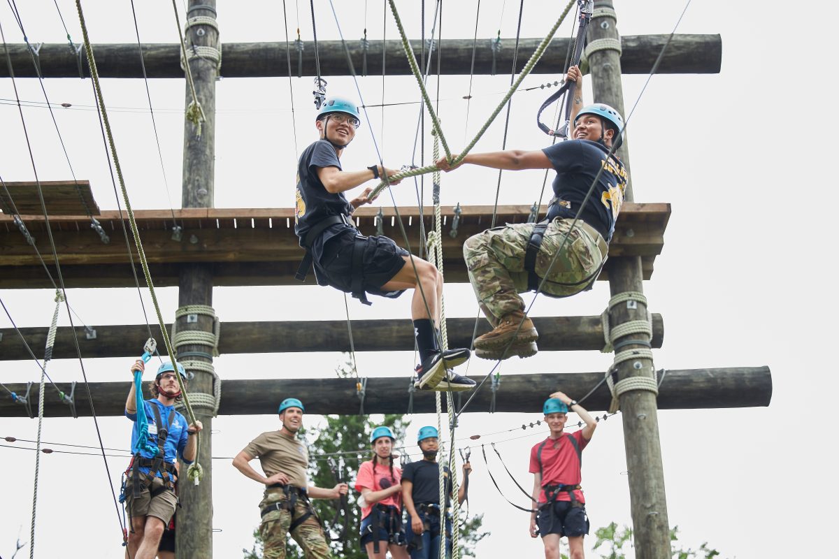 2 ROTC cadets traversing the high ropes course together, smiling with a group behind them cheering