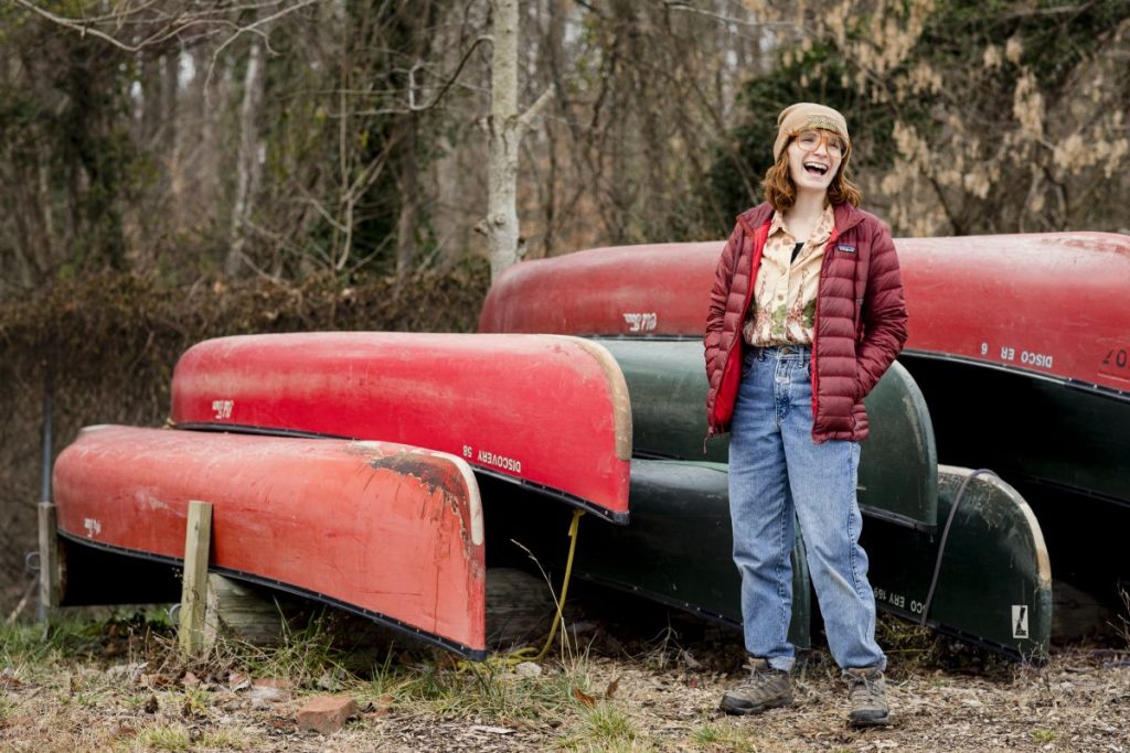 A photo of Outward Bound instructor Chloe wearing a puffy jacket, jeans, and hiking boots. She has a joyous smile on her face while posing in front of a stack of canoes. 