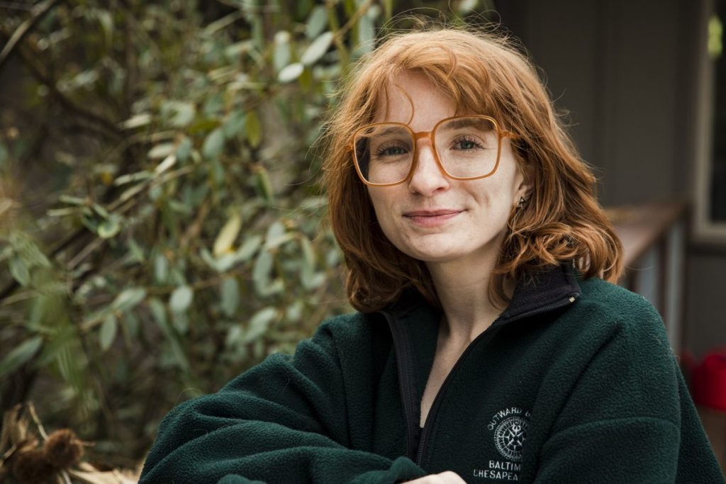 An up close portrait of Outward Bound instructor Chloe. She has a wavy red bob hair cut, and vintage, large glasses on. She is wearing a vintage Outward Bound fleece. She smiles warmly at the camera.
