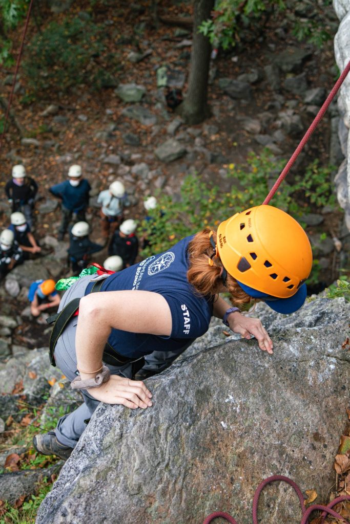 Outward Bound instructor Chloe is rock climbing in the forest. She wears a helmet and harness as she takes a big step up a bolder. Her students are on the ground looking up at her.