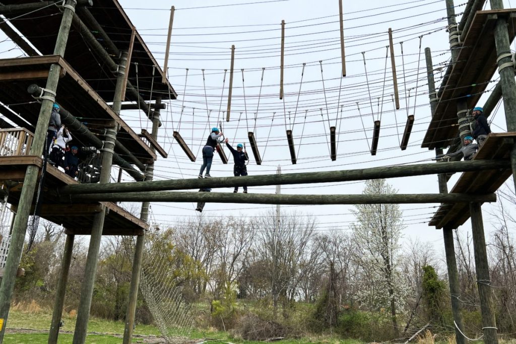 Two Outward Bound Board Members traverse the high-ropes course together and are high-fiving in the middle while others watch.